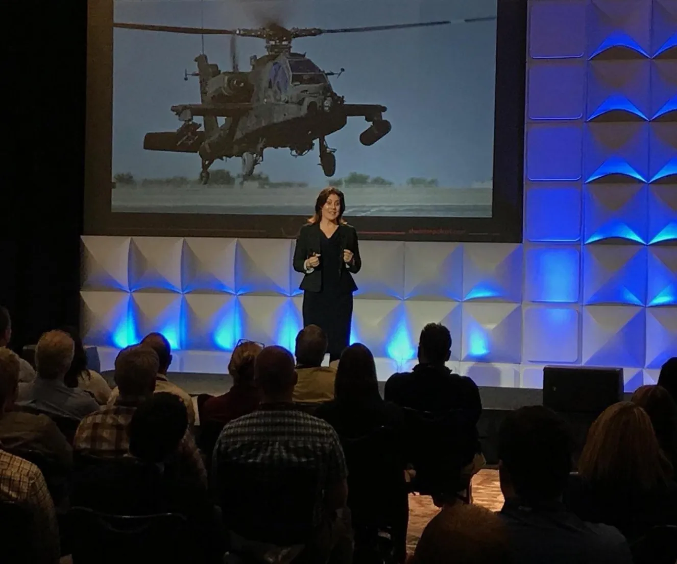 Shannon Huffman Polson on stage before a large audience, an Apache helicopter on the screen behind her