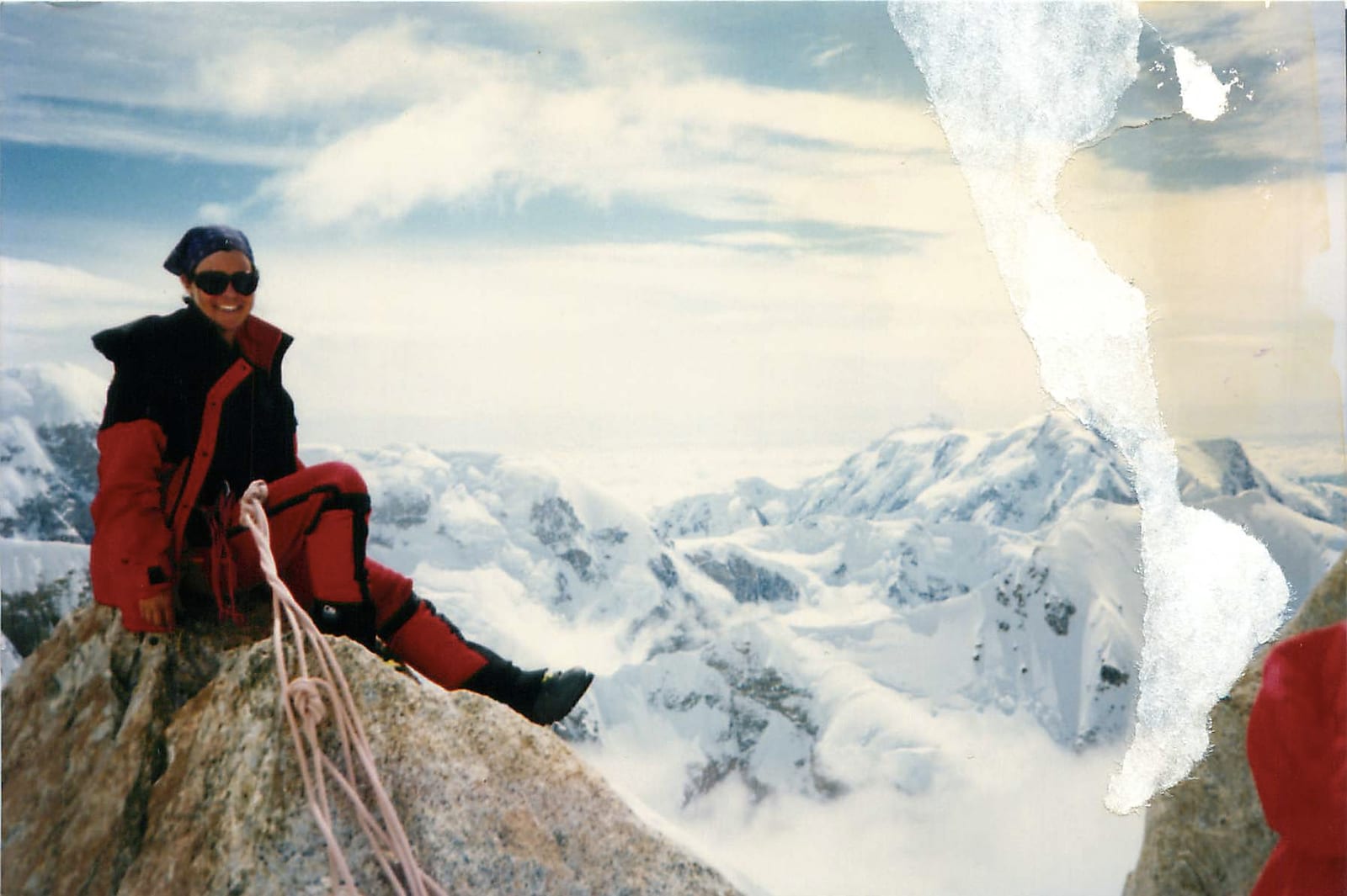 A climber rests on a rocky ridge above snow-covered peaks on Denali